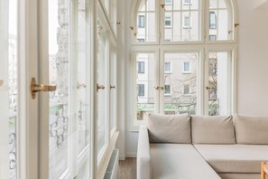 A room with a large window, a beige sofa, and a view of buildings outside. - Frankfurter Allee Deluxe Apartments by Sabine (Berlin)
