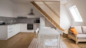 Eine moderne Küche mit weißen Schränken, einer Holztreppe, einer Essecke mit weißem Tisch und Stühlen sowie einem Sofa mit orangefarbenem Kissen.