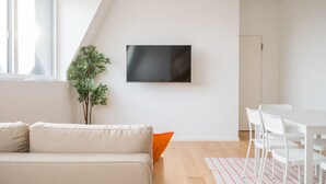 Ein modernes Wohnzimmer mit einer Couch, einem Esstisch mit Stühlen, einem an der Wand befestigten Fernseher und einer Zimmerpflanze.