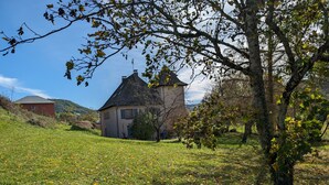 Une maison avec un toit en pente, entourée d’un champ verdoyant parsemé de fleurs jaunes, et un arbre aux feuilles vertes au premier plan.
