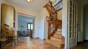Un hall d’entrée spacieux, doté d’un escalier en bois, d’un sol carrelé et donnant sur un salon avec un canapé et une lampe.