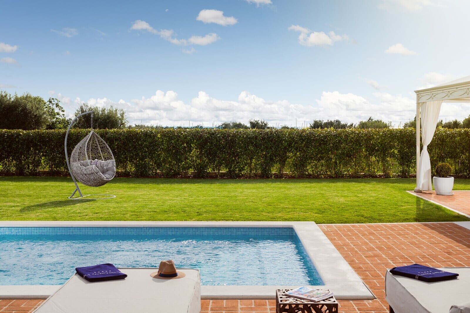A swimming pool with lounge chairs, a hanging chair, and a pergola with white curtains.