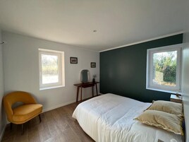 Un dormitorio con una cama, una silla, una mesita y dos ventanas.