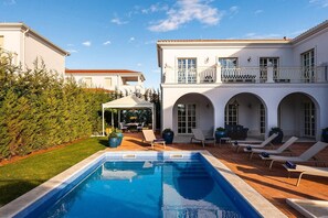 En pool med liggestole, en overdækket terrasse og en bygning med buede vinduer og en balkon.
