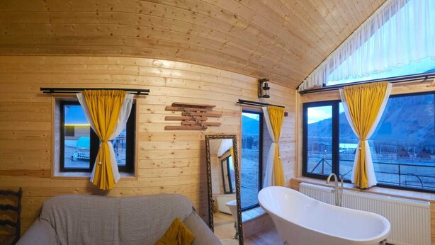 A cabin with wooden walls, a bathtub, a sofa, and large windows with curtains. - Mood villa kazbegi is city centre of Stepantsminda, with mountimes view. For you (Stepantsminda)