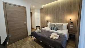 A modern hotel room with a wooden headboard, a large bed with a dark bedspread, two bedside lamps, and a wooden floor. - ARYA Cagliari (Cagliari)