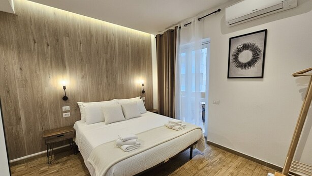 A modern hotel room with a wooden accent wall, a large bed, bedside lamps, a framed artwork, and a window with curtains. - ARYA Cagliari (Cagliari)