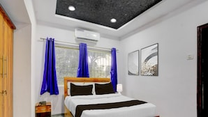 A bedroom with a wooden bed, a nightstand, a window with curtains, an air conditioning unit, and two wall-mounted artworks. - Deluxe Seven Hills Aurangabad (Chhatrapati Sambhajinagar)