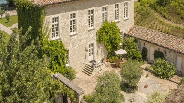 Edificio de piedra con tejado de baldosas, rodeado de vegetación y un pequeño patio.