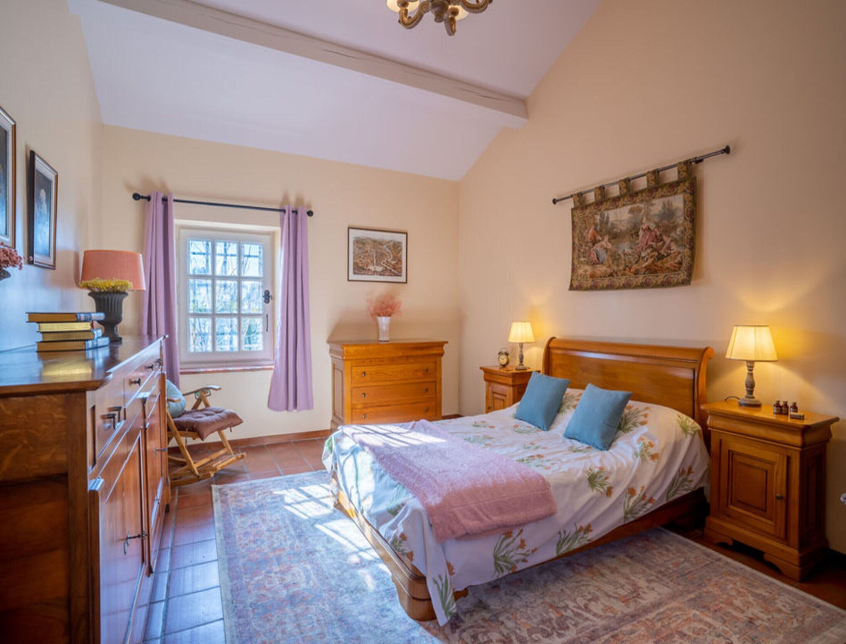 A bedroom with a bed, bedside tables, a dresser, a window with curtains, a painting on the wall, and a wooden chair.