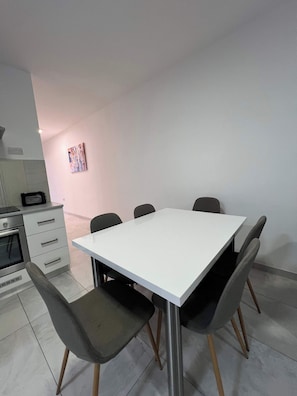 A modern dining area with a white table, six chairs, a kitchen with a microwave, and a small painting on the wall. - Swieqi Modern 3 BDR Flat by PMI Malta (Is-Swieqi)