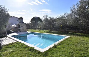 Una piscina rectangular con borde de piedra, rodeada por un césped y árboles.