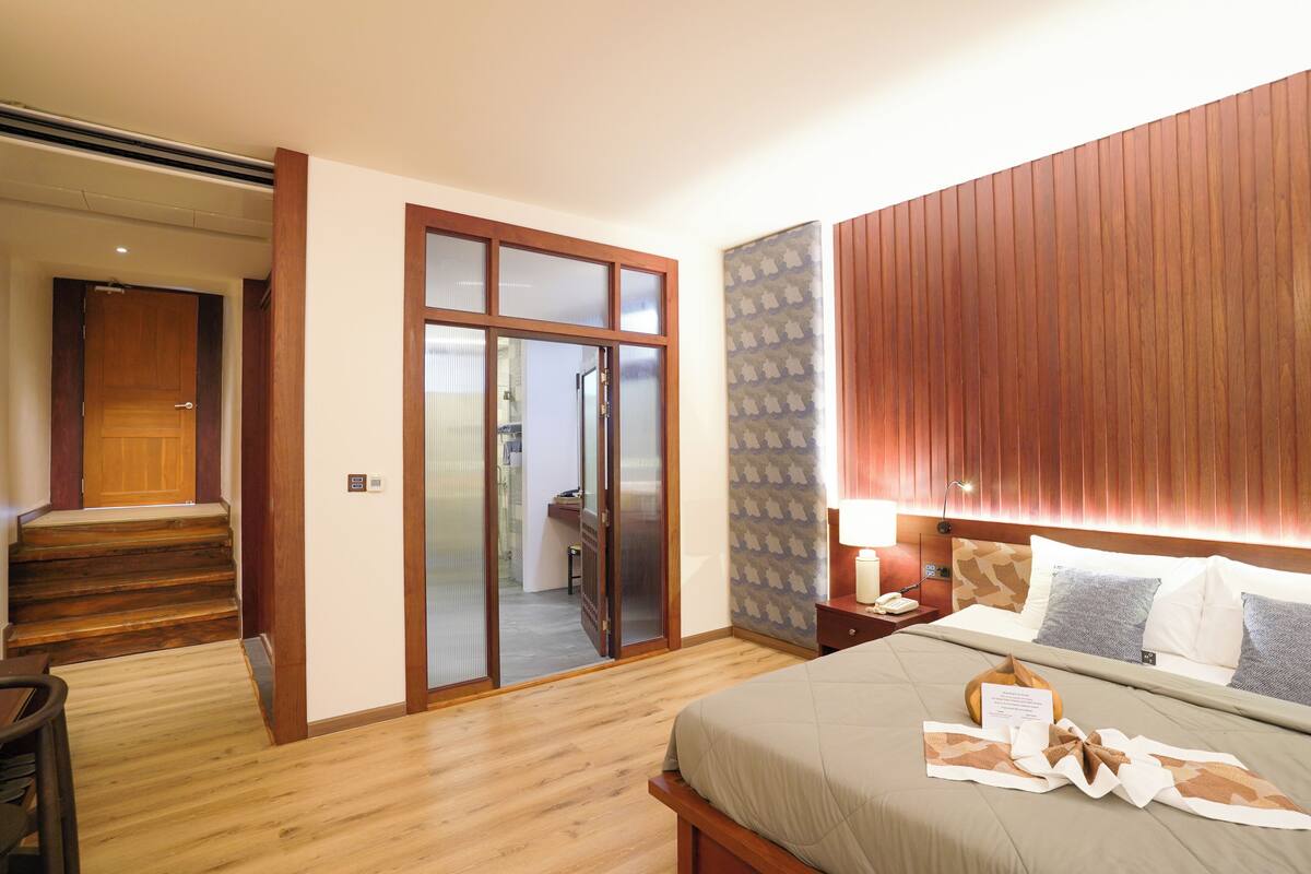 A hotel room with a wooden floor, a bed with a beige bedspread, a nightstand with a lamp, and a sliding door leading to a bathroom.
