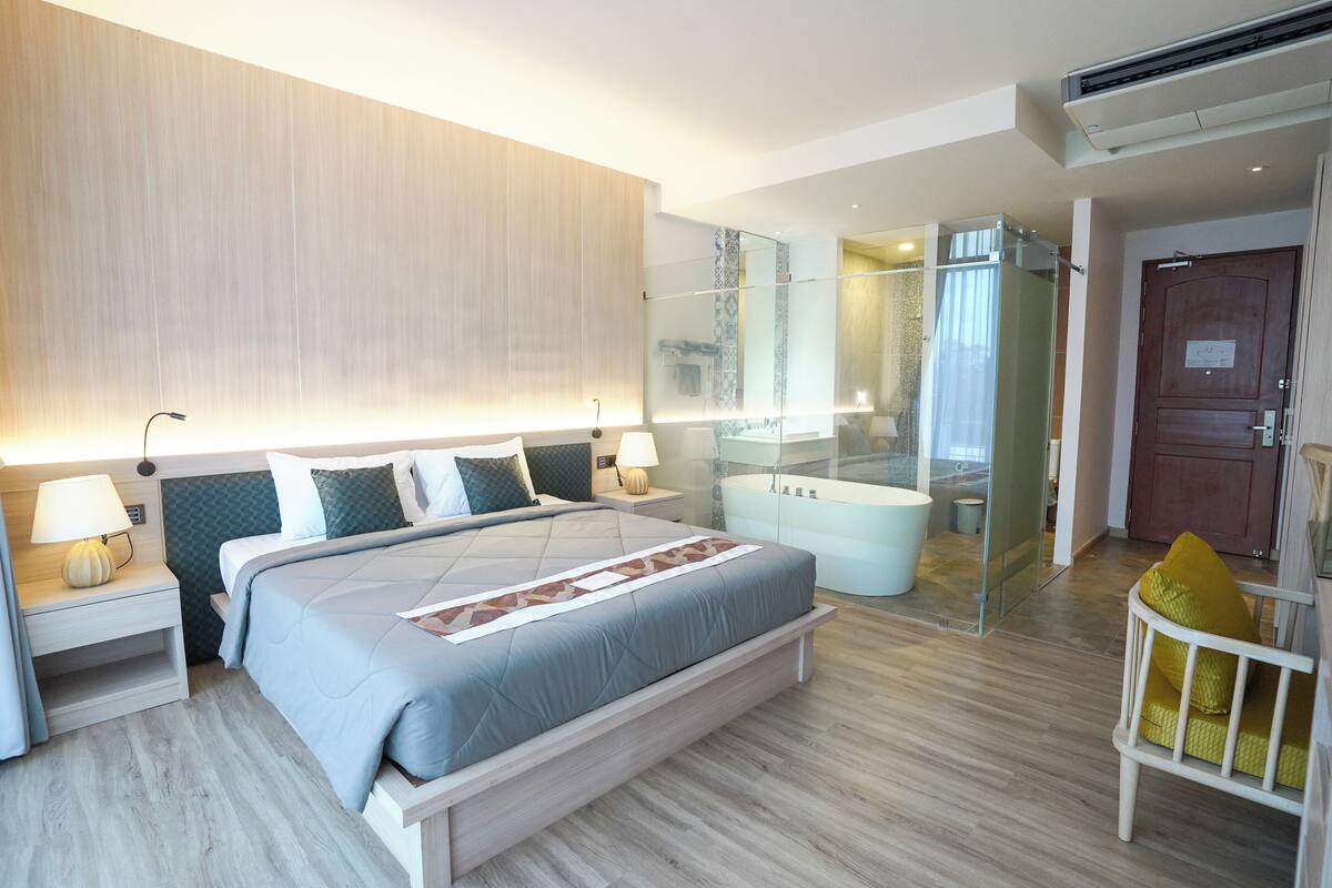 Moderni hotellihuone, jossa on suuri sänky, yöpöydät lampuineen, lasiseinällä varustettu suihku ja puuovi.