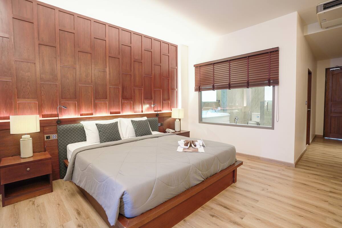 A hotel room with a large bed, wooden headboard, bedside tables with lamps, a window with blinds, and a wooden floor.