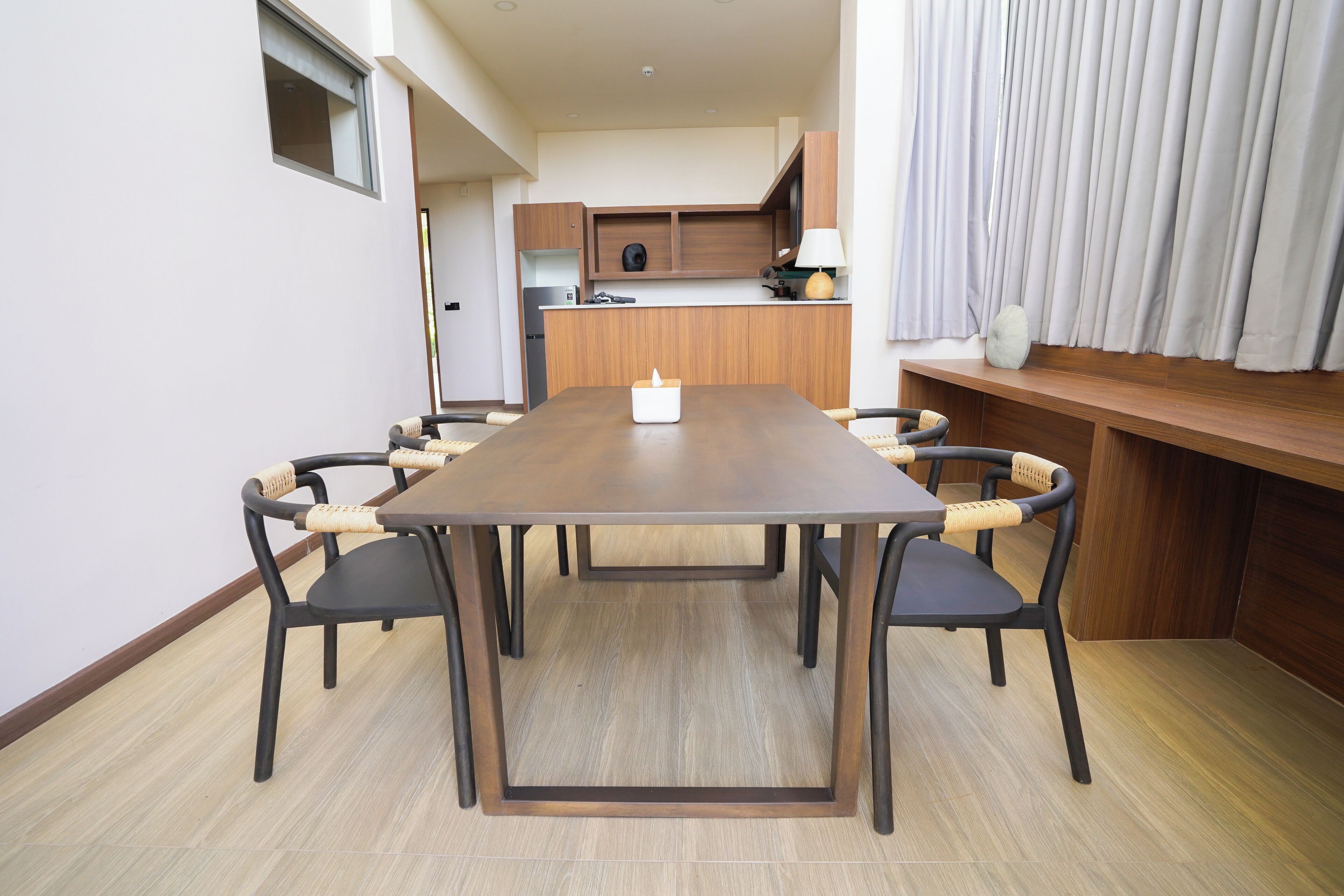 A dining area with a wooden table and chairs, a kitchen in the background, and a window with curtains.