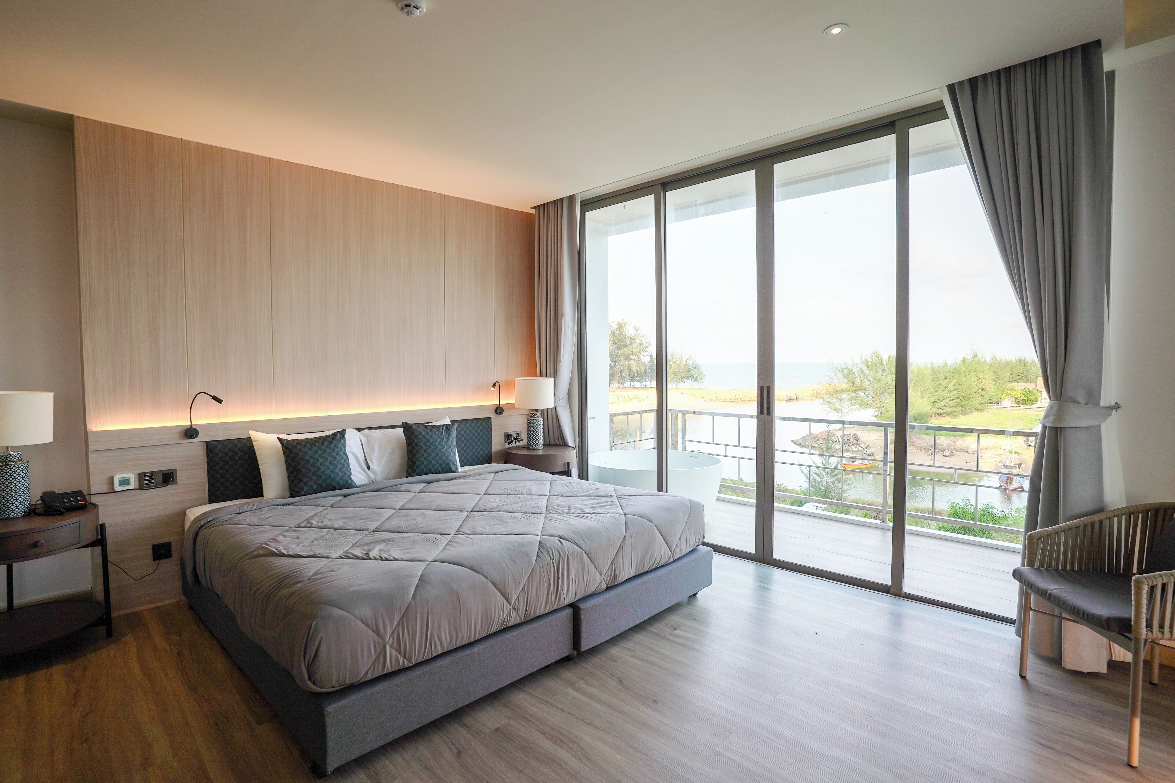 A modern hotel room with a large bed, bedside tables, a view of the outdoors, and a balcony with a railing.