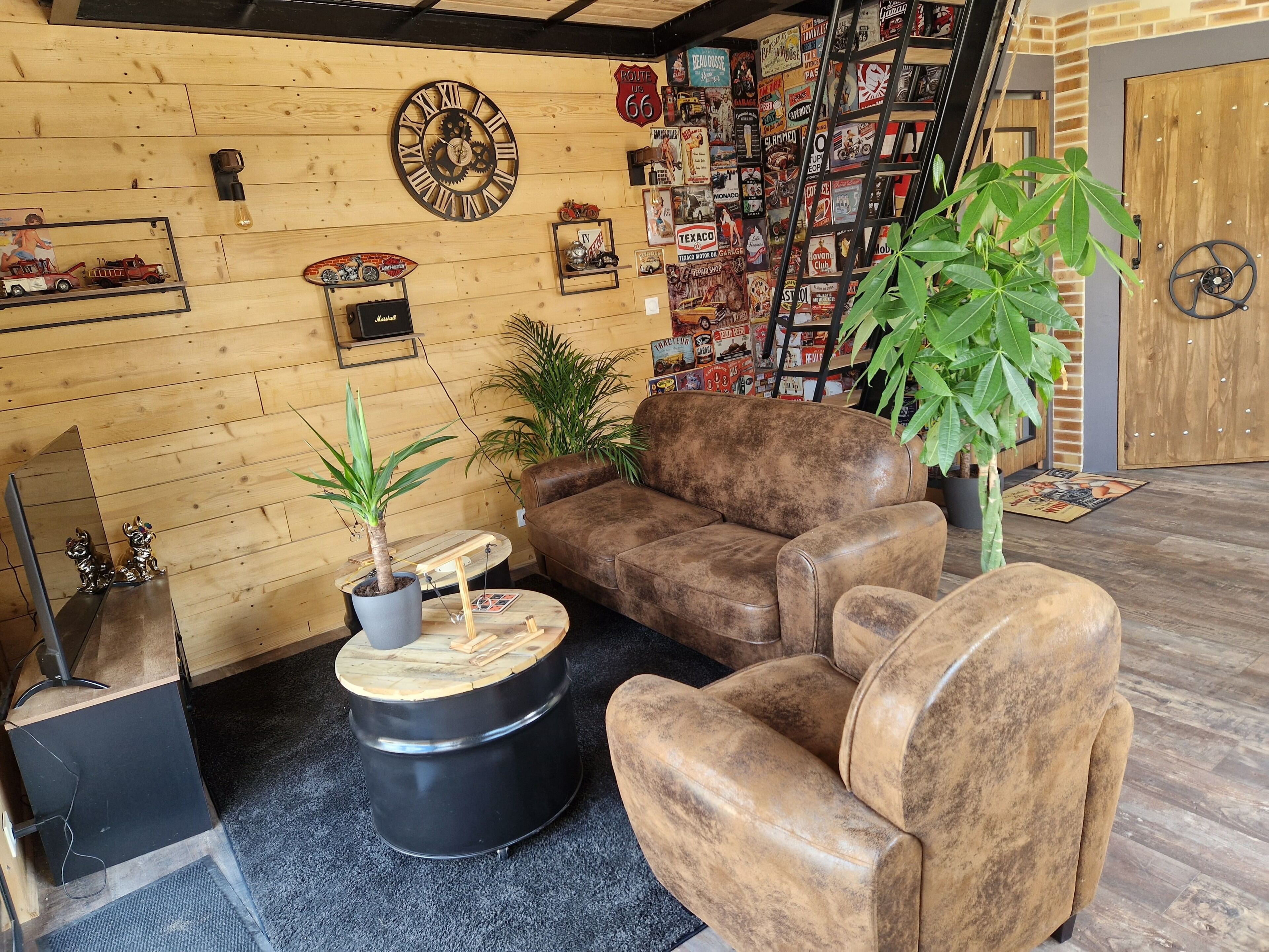 Ein gemütliches Zimmer mit einer Holzwand, einem braunen Ledersofa, einem runden schwarzen Hocker und einer Topfpflanze.