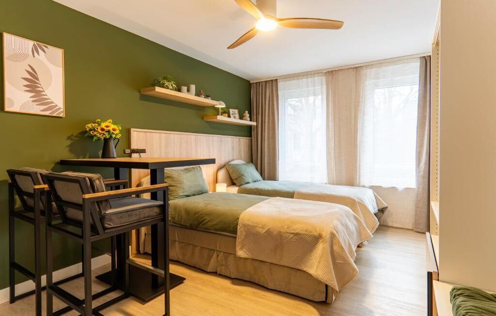 A room with a bed, a desk with chairs, a ceiling fan, and a window with curtains.
