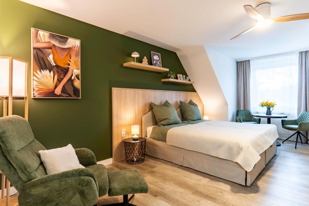 Una habitación de hotel moderna con una cama, un sillón verde, una mesita pequeña y un cuadro en la pared.
