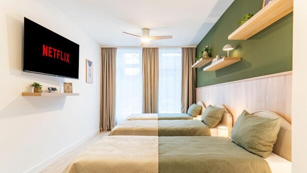 A hotel room with a TV displaying the Netflix logo, a ceiling fan, two beds with matching bedding, a window with curtains, and a shelf with decorative items. - be Apart Hotel Serviced Apartments (Hannover)
