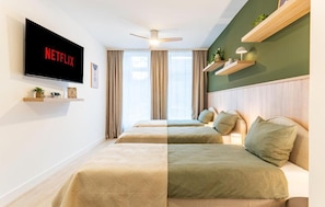 A hotel room with a TV displaying the Netflix logo, a ceiling fan, two beds with matching bedding, a window with curtains, and a shelf with decorative items.