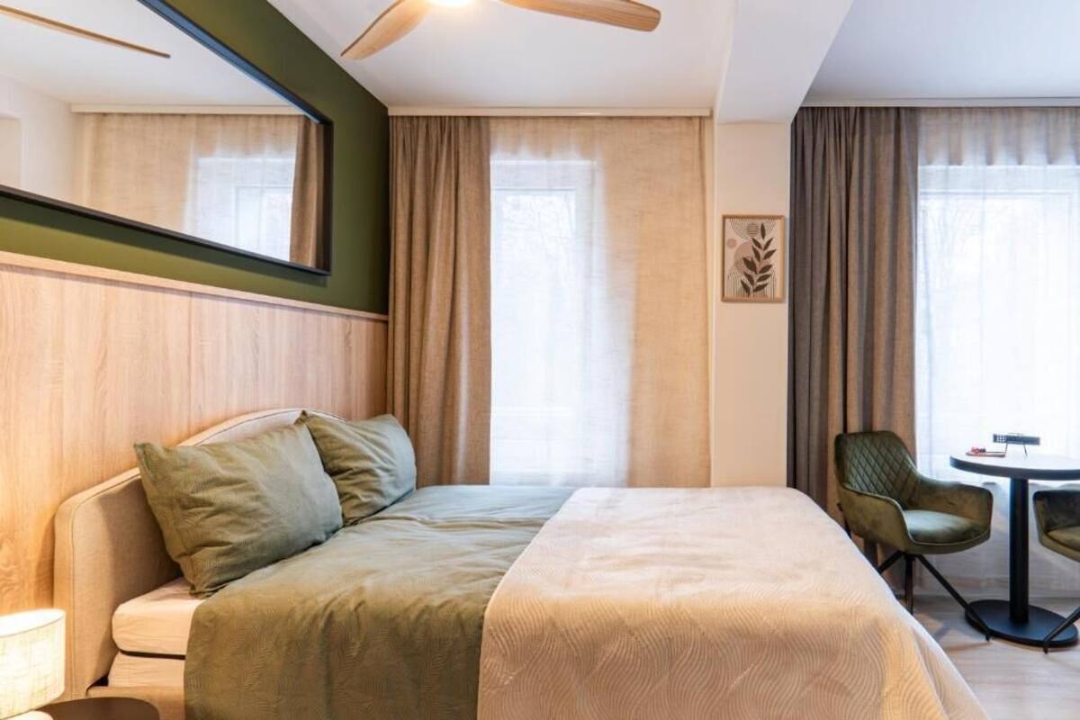 Habitación de hotel con cama, una silla, una mesita, espejo y ventana con cortinas.