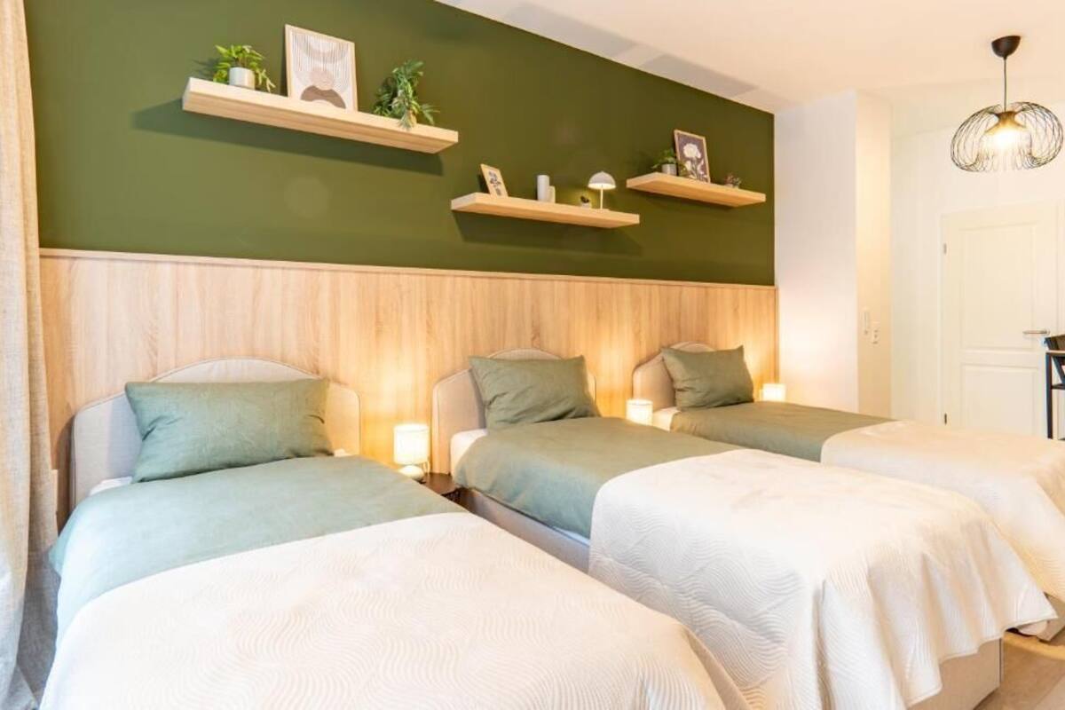 Habitación de hotel con tres camas, cabecero de madera, paredes verdes y estantes con plantas y fotografías.