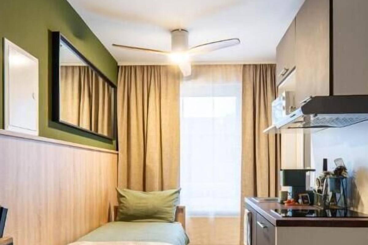 Habitación de hotel compacta con kitchenette, dos camas, ventilador de techo y ventana con cortinas.