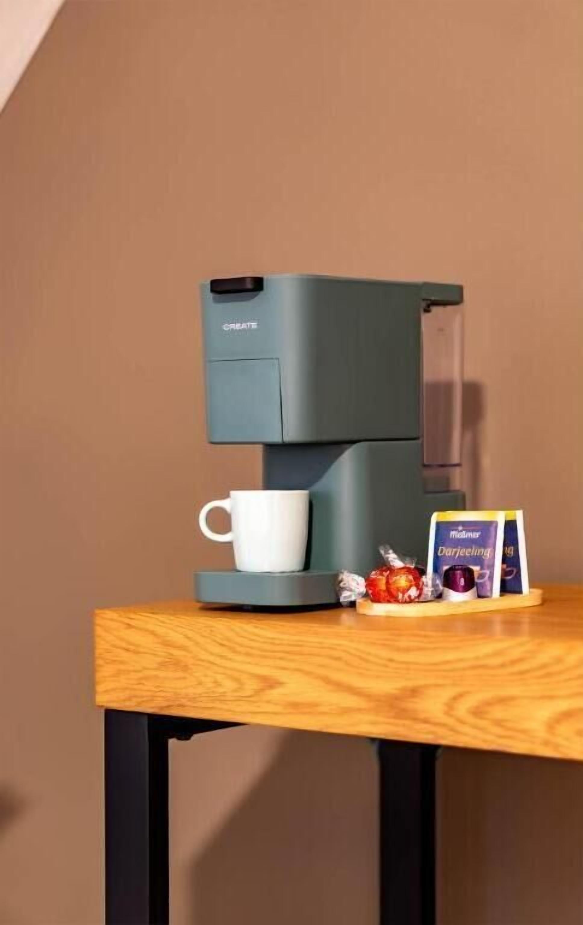 Una cafetera sobre una mesa de madera, con una taza blanca y una caja de cápsulas de café.