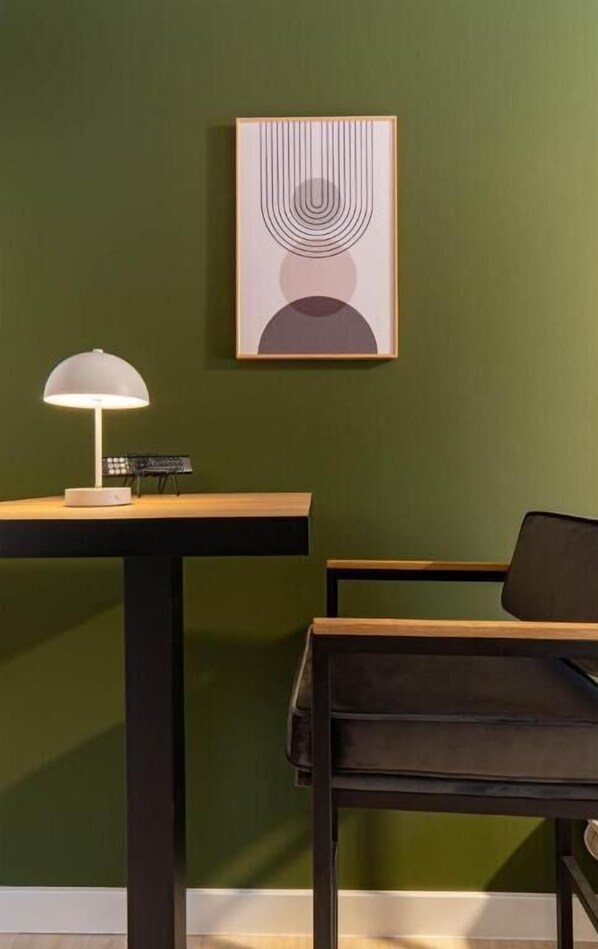 A room with a green wall, a framed abstract artwork, a desk with a lamp, and a chair.