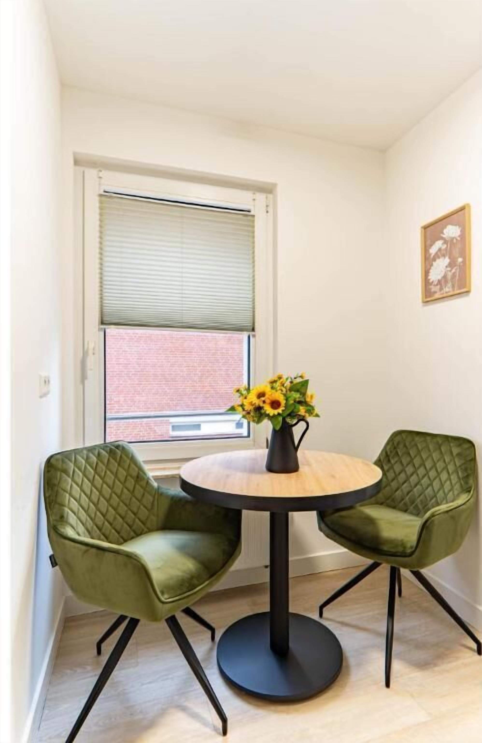 Quarto de canto com mesa redonda, duas cadeiras verdes, um vaso com flores, janela com persianas e um quadro na parede.