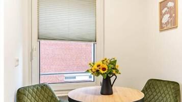 A corner room with a round table, two green chairs, a vase with flowers, a window with blinds, and a picture on the wall.
