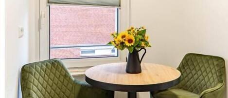 Quarto de canto com mesa redonda, duas cadeiras verdes, um vaso com flores, janela com persianas e um quadro na parede.