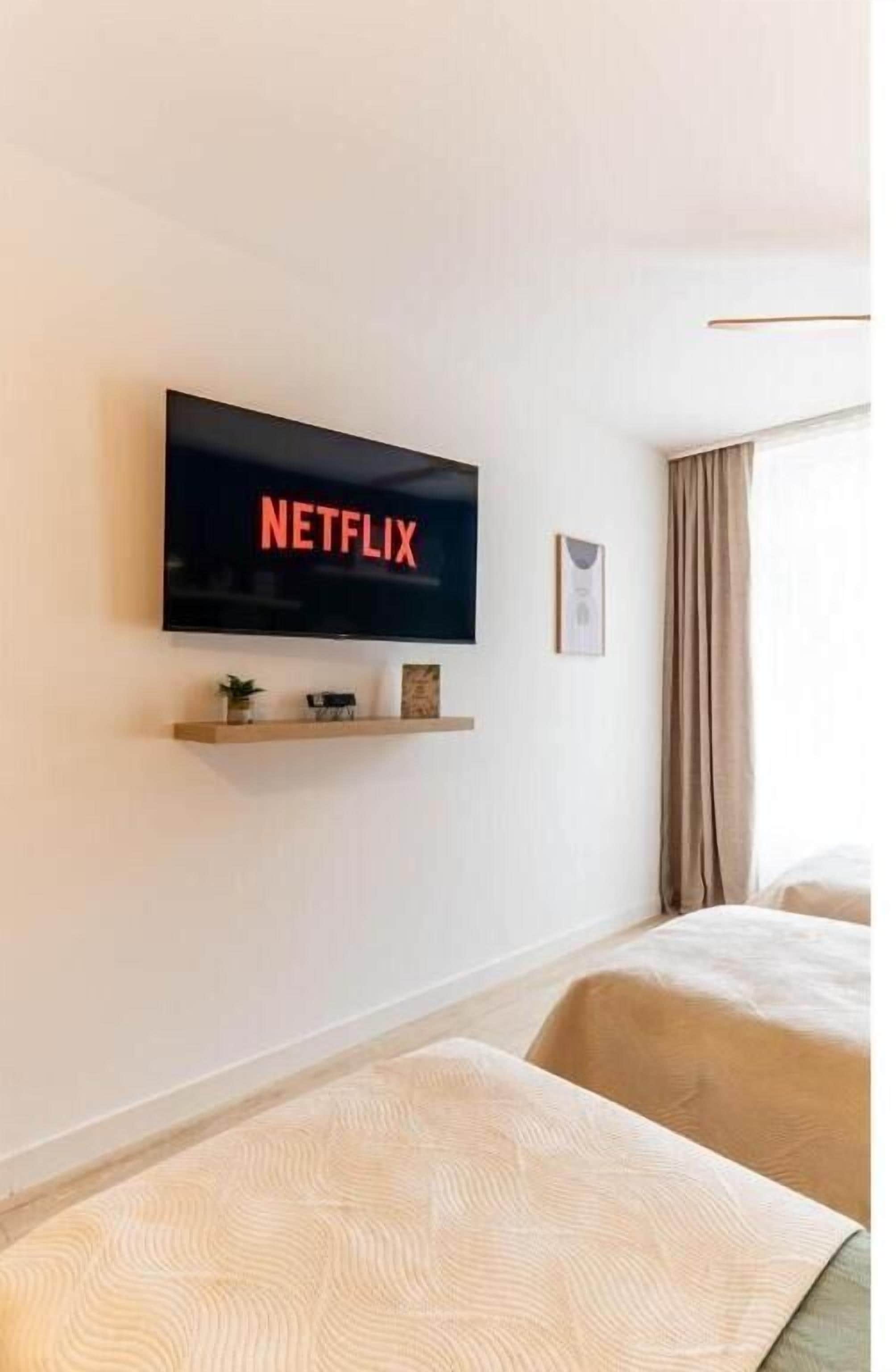 A modern hotel room with a mounted TV displaying 'NETFLIX,' a small shelf with decorative items, two beds with beige bedding, and a large window with curtains.