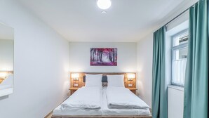 A neatly made bed with white linens, a painting of a forest on the wall, and a window with curtains.