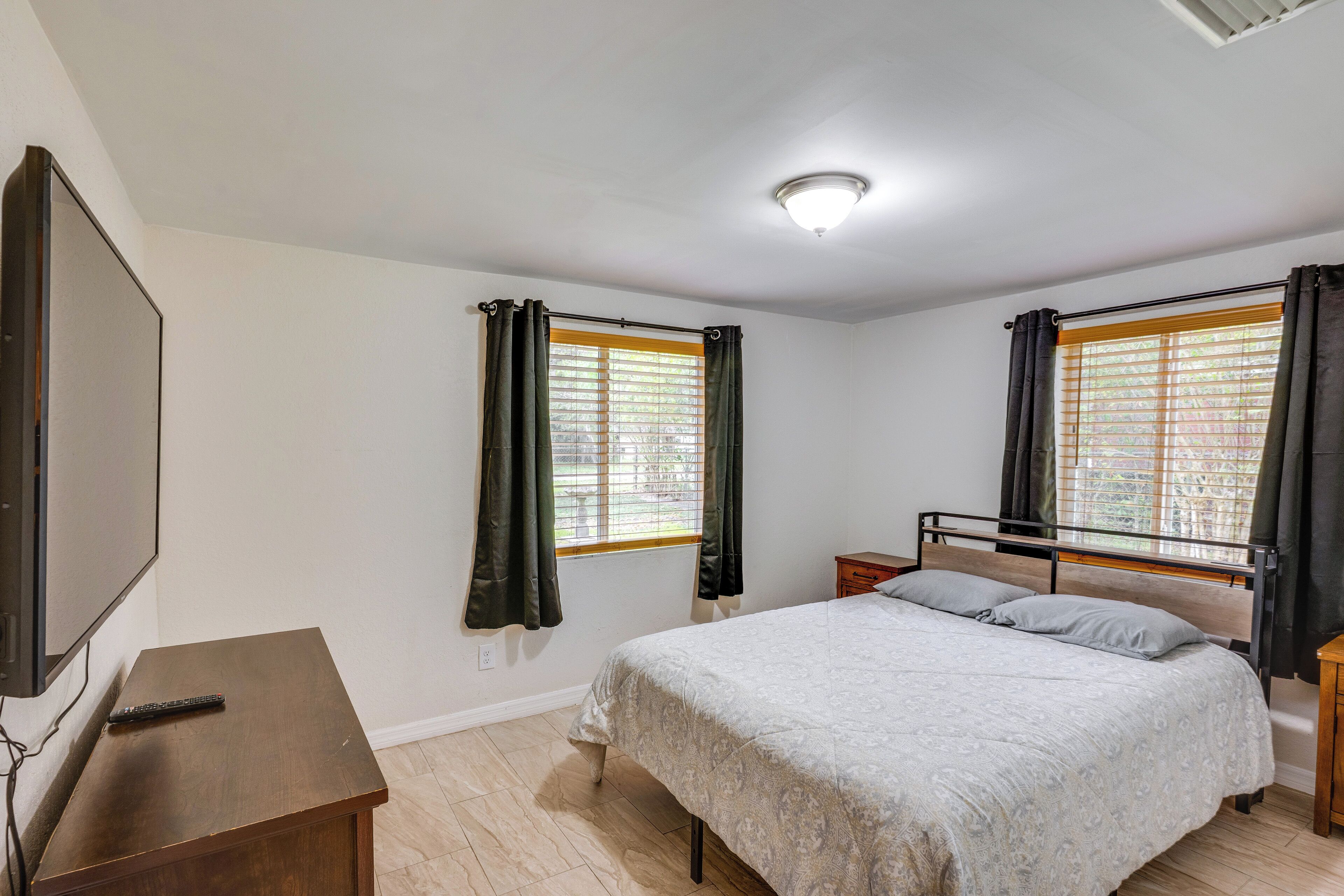 Une chambre à coucher avec un lit, un téléviseur posé sur un meuble, deux fenêtres avec des rideaux et un luminaire au plafond.
