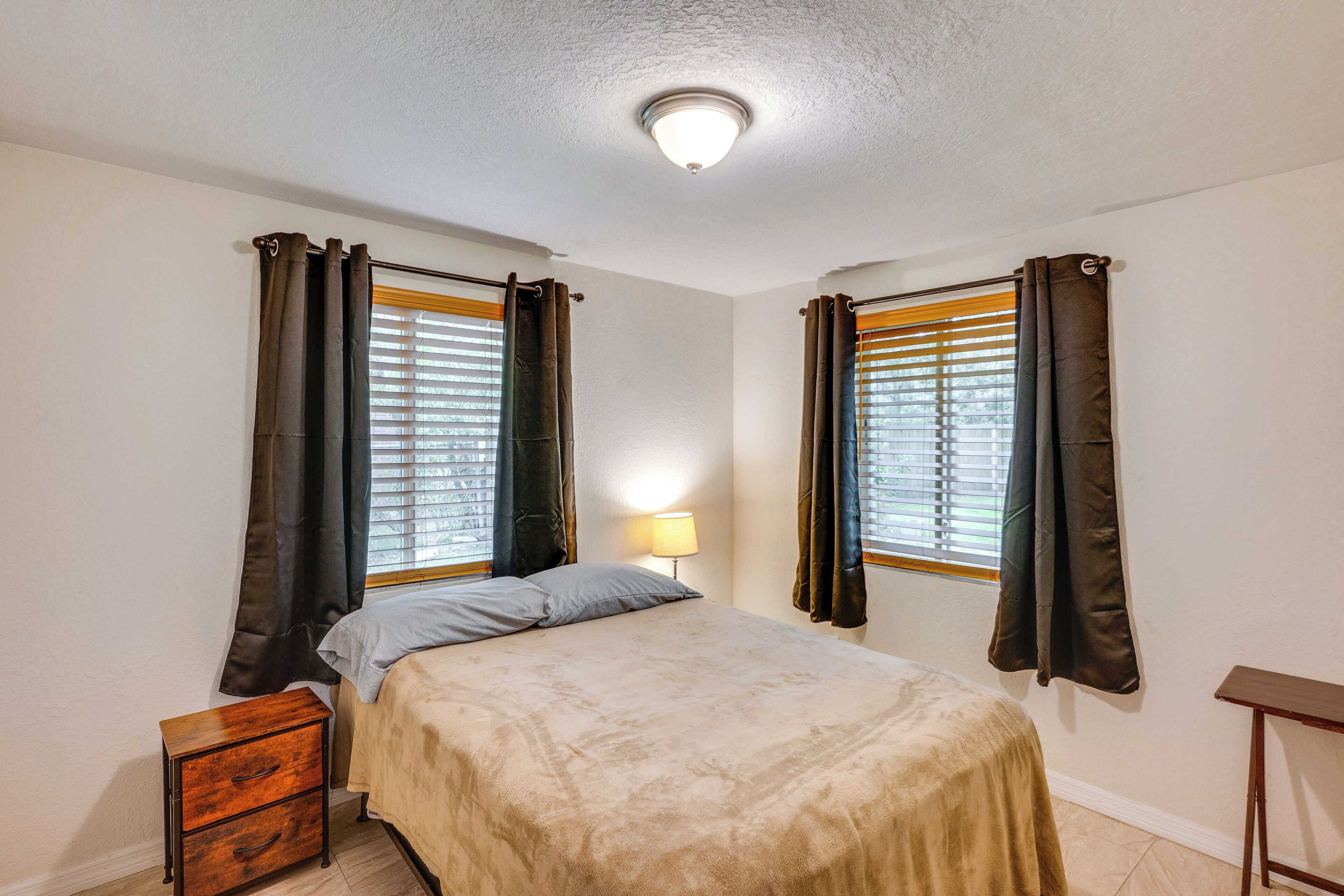 Une chambre à coucher avec un lit, deux fenêtres avec des rideaux, une table de chevet et une lampe.