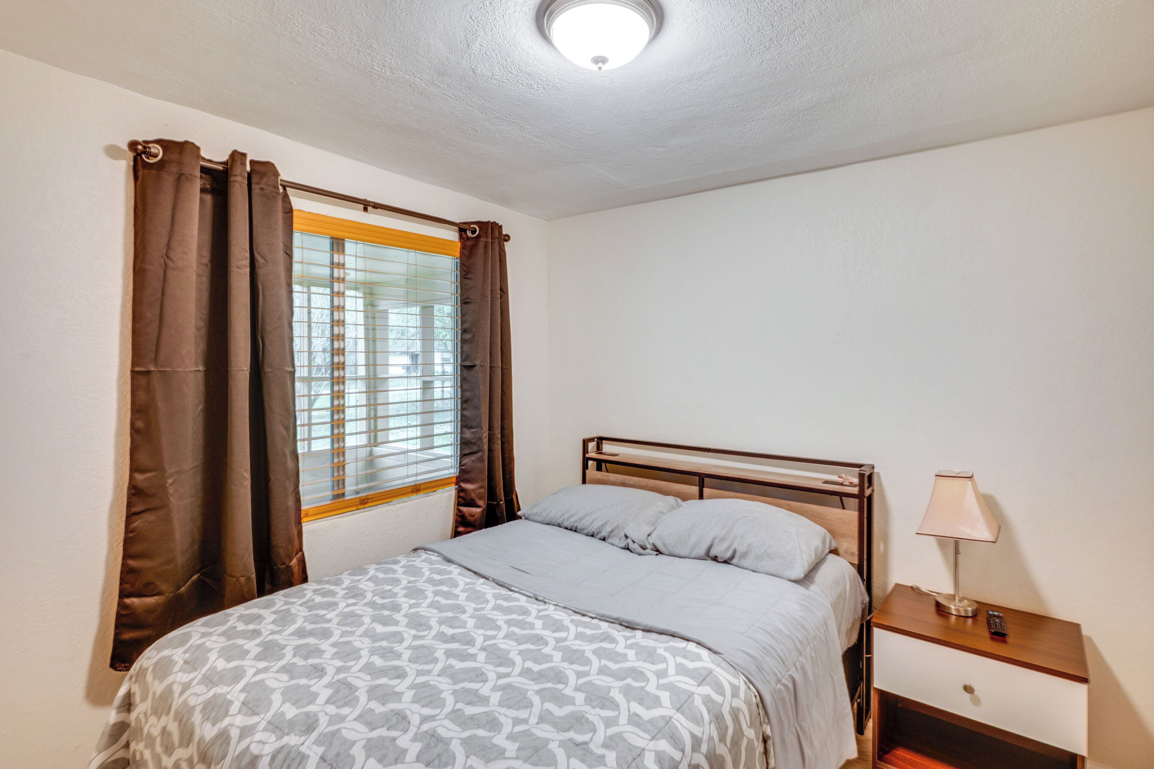 Un dormitorio con cama, cortinas, una ventana con persianas, una mesita de noche con lámpara y un aparador.