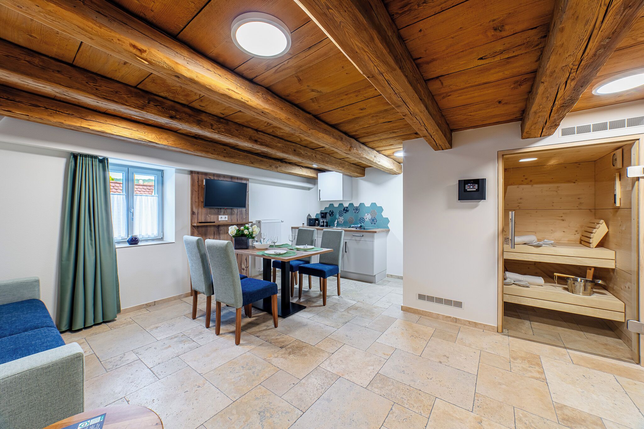 A room with a wooden ceiling, a dining area with chairs and a table, a kitchen with cabinets and a TV, and a sauna area.