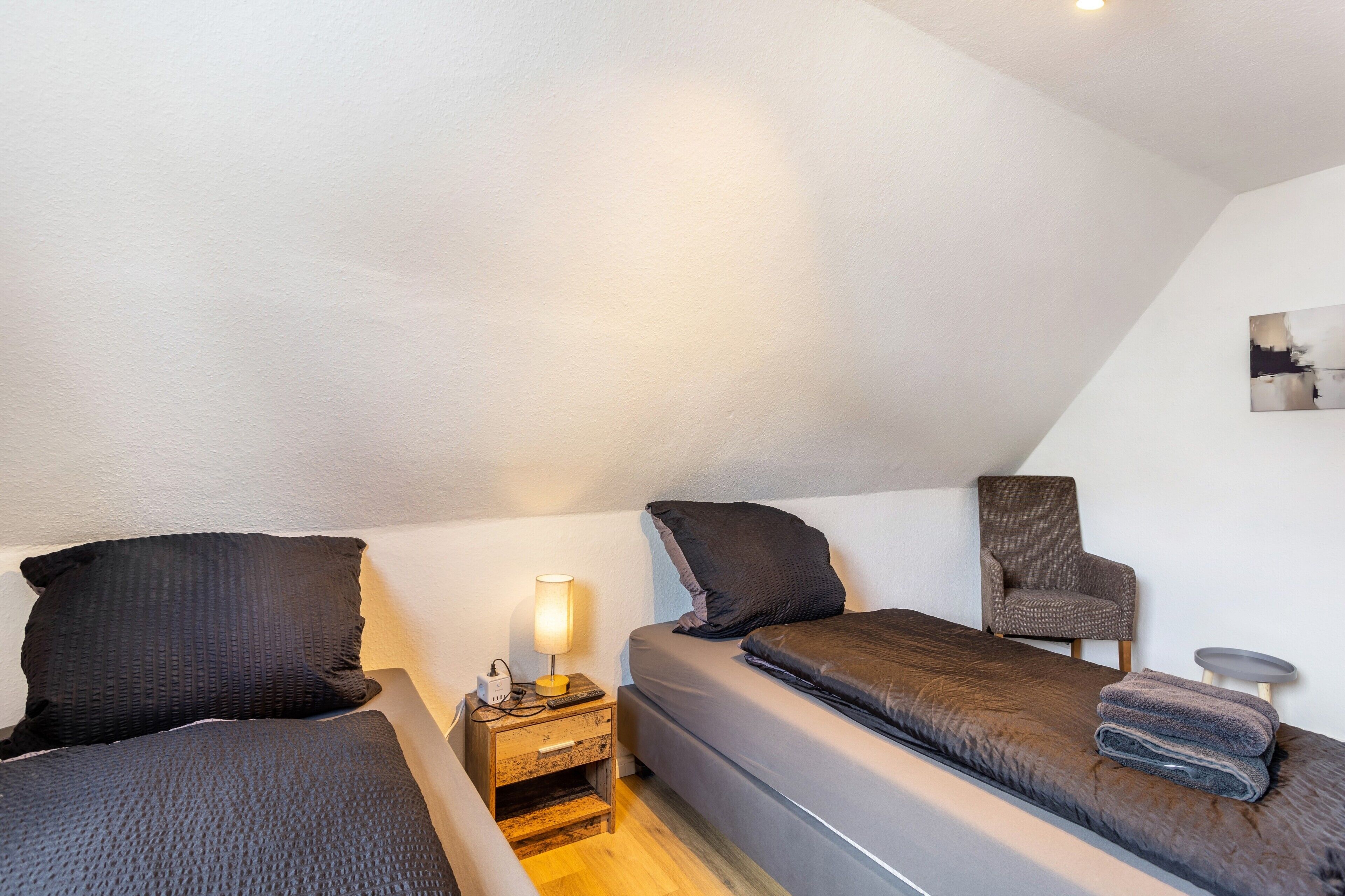 Una habitación pequeña y acogedora con dos camas, una silla, una mesita de noche con lámpara y una pequeña mesa.