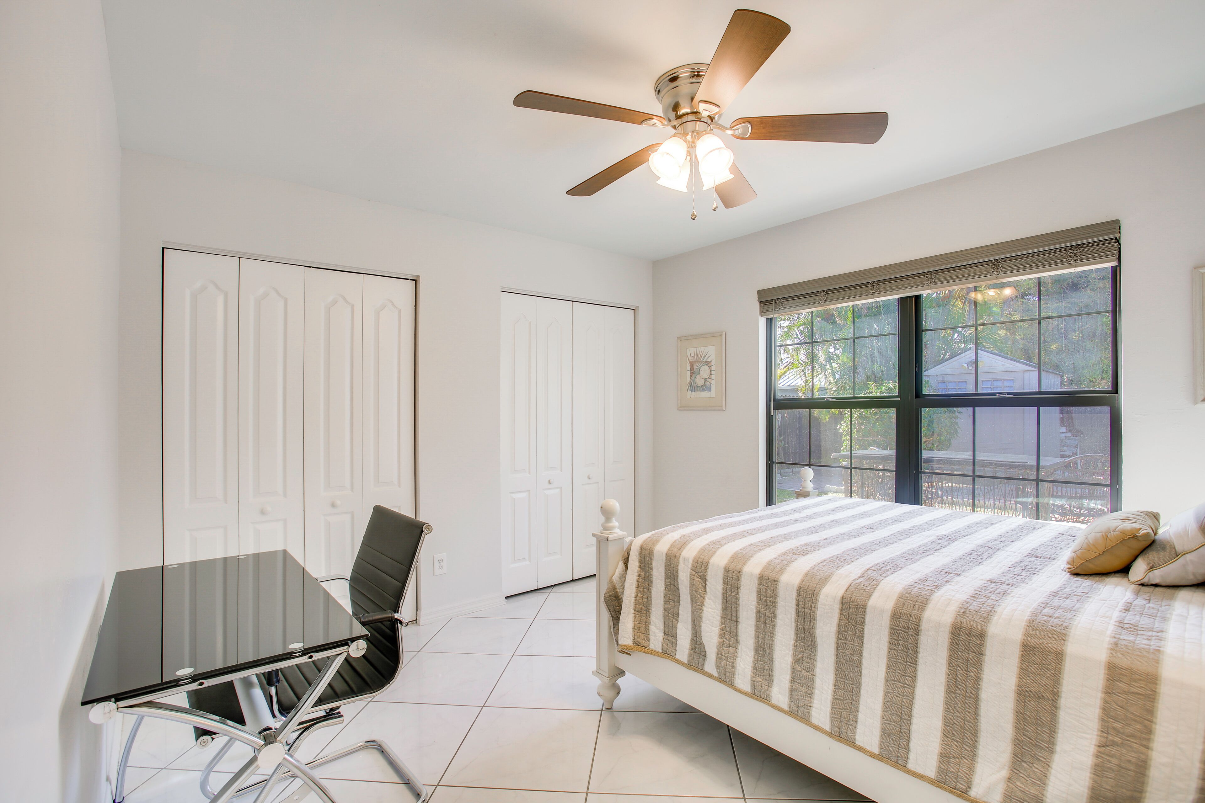 A bedroom with a bed, a desk, a chair, a ceiling fan, and a window with a view.