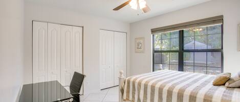 A bedroom with a bed, a desk, a chair, a ceiling fan, and a window with a view.