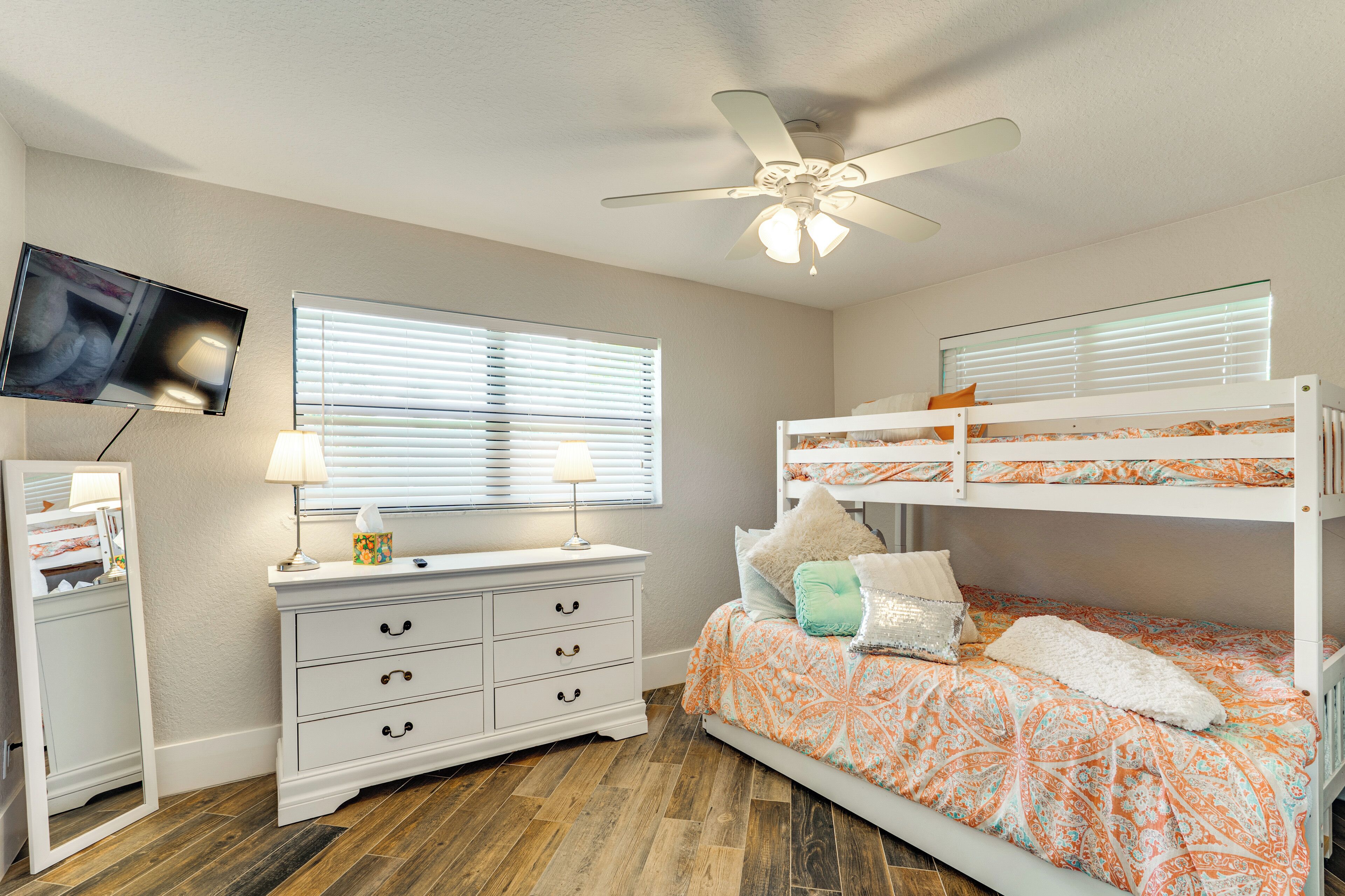 A bedroom with a bunk bed, a dresser, a television mounted on the wall, and a ceiling fan.