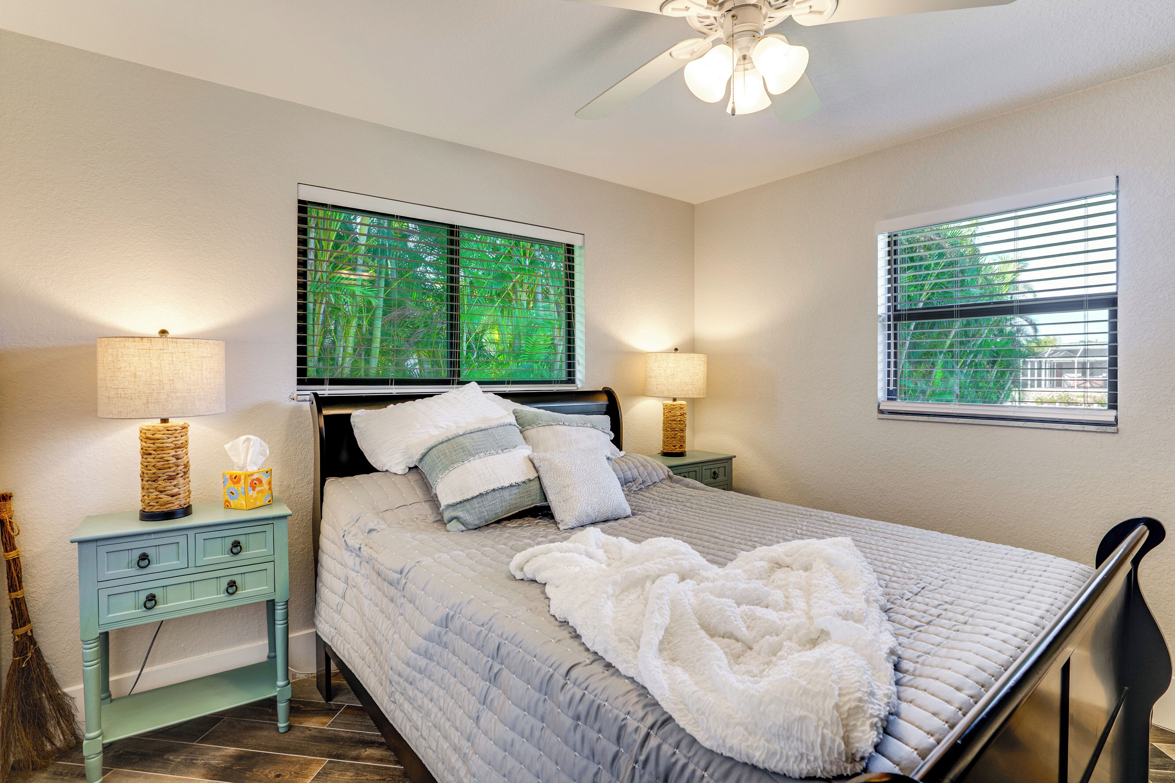 A bedroom with a bed, two bedside tables, a ceiling fan, and two windows with blinds.