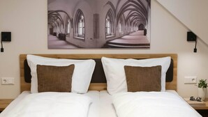 A hotel room with a large bed, two pillows, a headboard, a wall-mounted picture, bedside lamps, and a small plant on a nightstand.