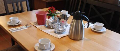 Een eettafel gedekt met kopjes, schoteltjes, bestek, een thermoskan en een bloemstuk.