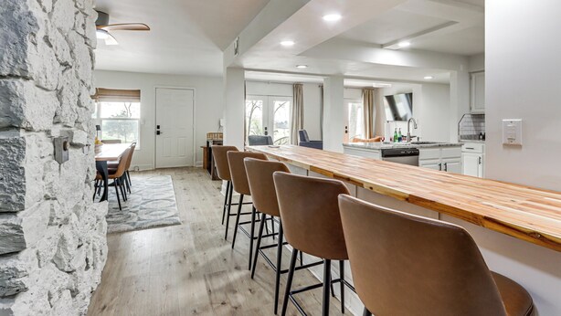 A modern kitchen with a long wooden bar, stone wall, and a dining area with chairs. - Patio + Golf Course Views: Monkey Island Escape! (Afton)