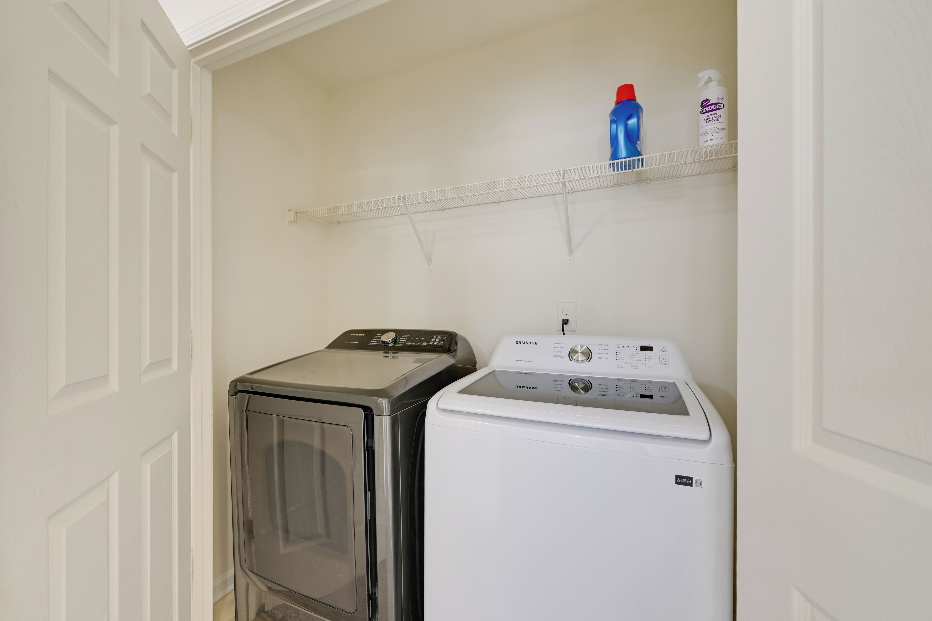 Uma área de lavanderia com máquina de lavar e secadora frontais, uma prateleira com produtos de limpeza e uma porta branca.
