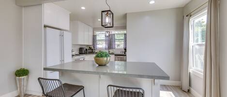 A modern kitchen with a central island, two bar stools, a potted plant, and a hanging light fixture.